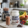 AtLast! Chocolate Milkshake meal replacement protein powder on kitchen counter with blended chocolate shake, bananas, apples, and berries