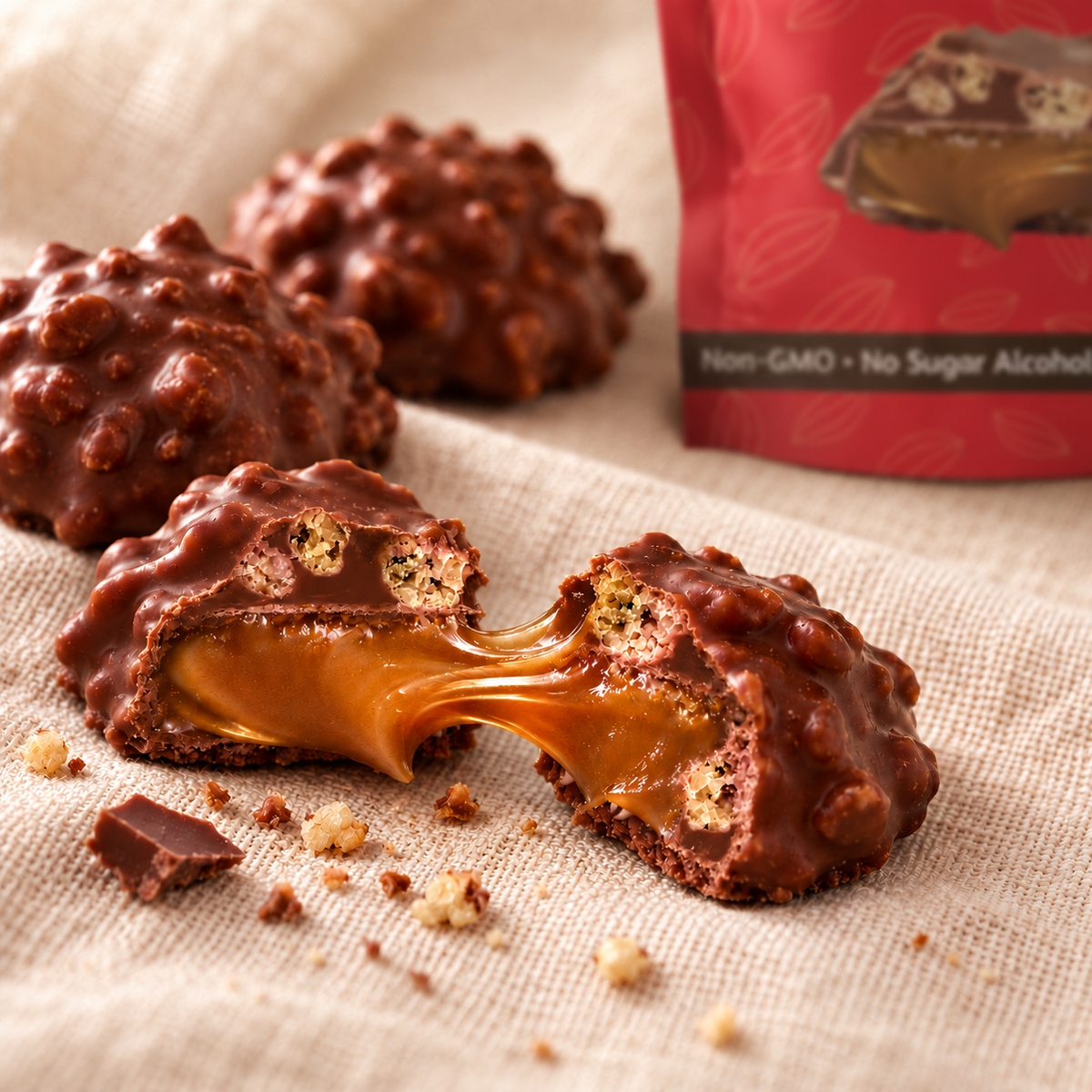 A milk chocolate crispy caramel broken in half, soft caramel stretching between the pieces, with two whole candies and a red candy pouch on a cream linen cloth.
